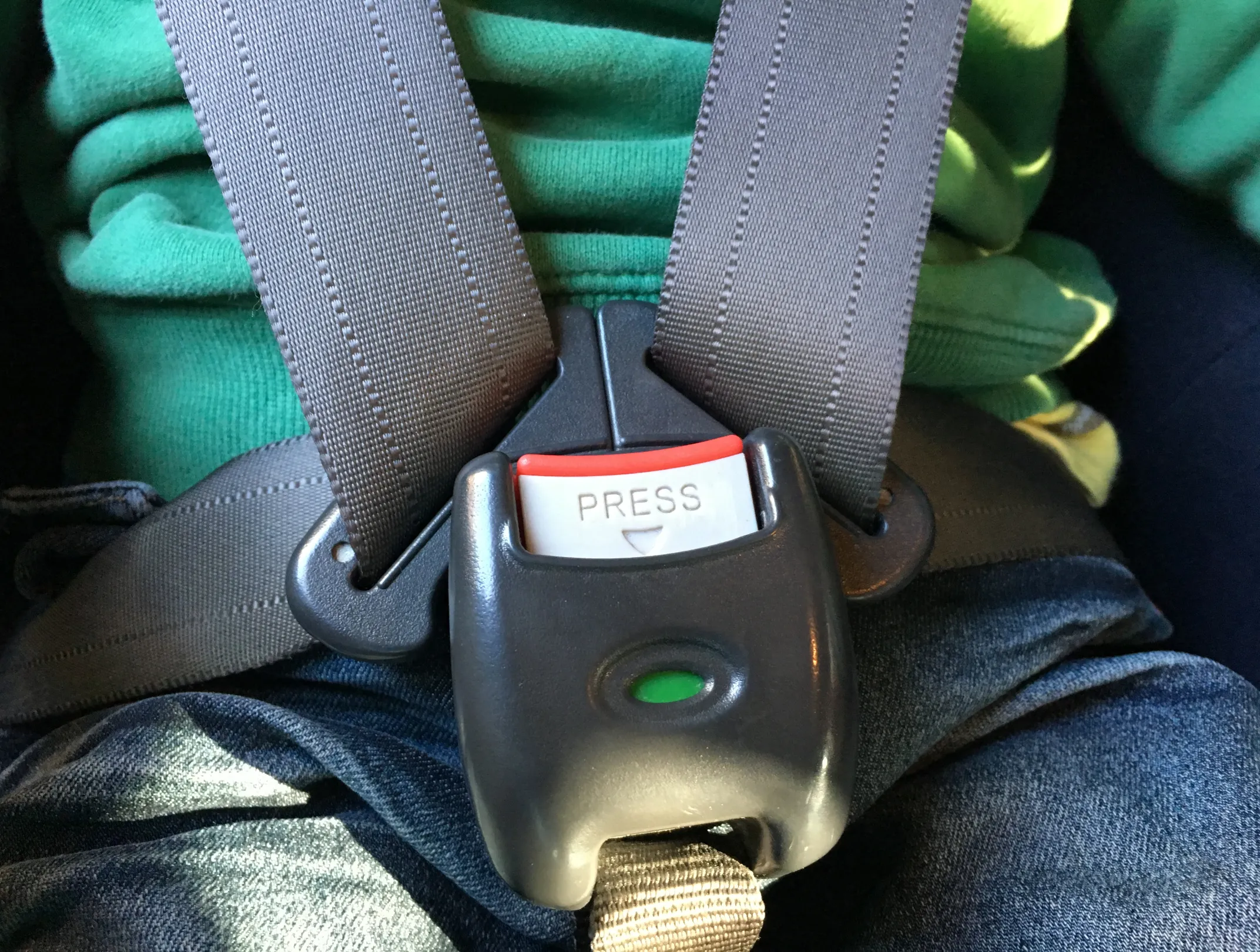 A child seated in a harness-type restraint, with grey webbing straps meeting at a central buckle marked PRESS across the chest and lap.