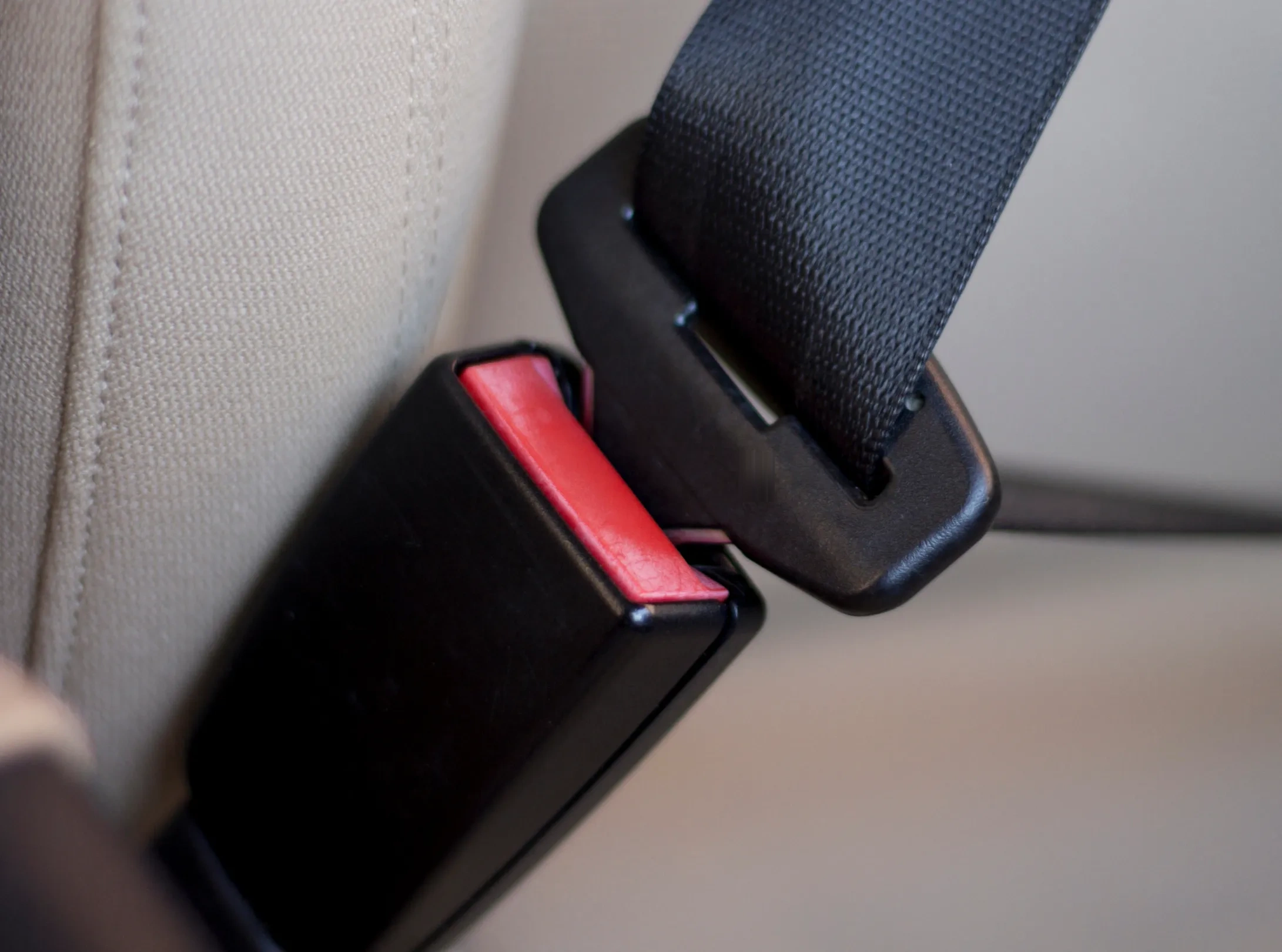 A seat belt tongue about to be inserted into a black buckle stalk with a red release button, photographed against a light vehicle seat.