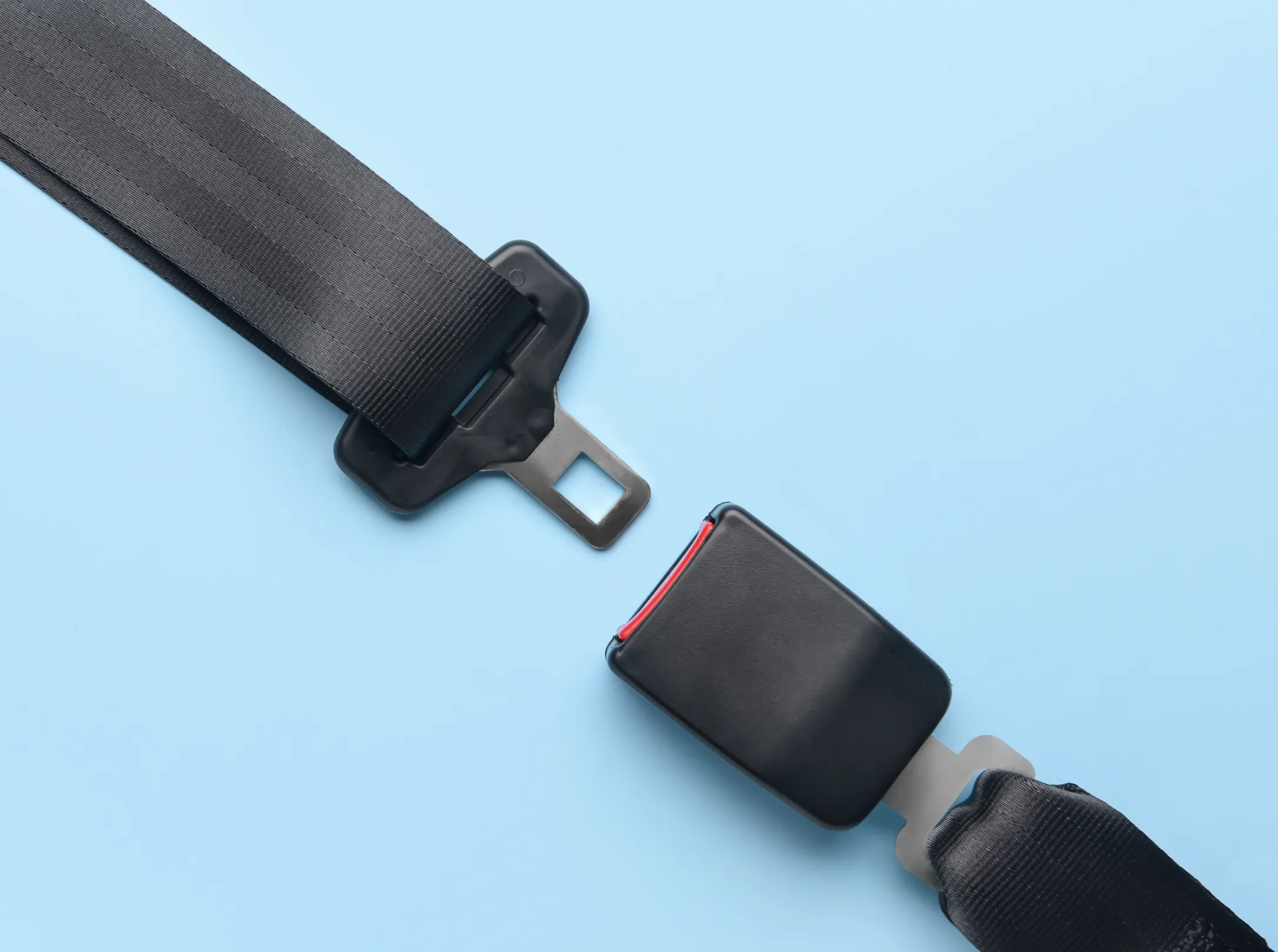 A seat belt tongue plate and buckle laid out separately on a plain pale background, showing the webbing, latch plate and release button.